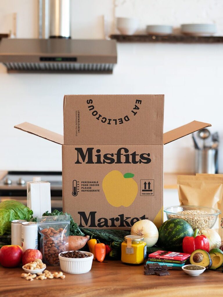 A box labeled "Misfits Market" is on a kitchen counter, surrounded by assorted fruits, vegetables, nuts, and pantry items. The kitchen background includes shelves and appliances.