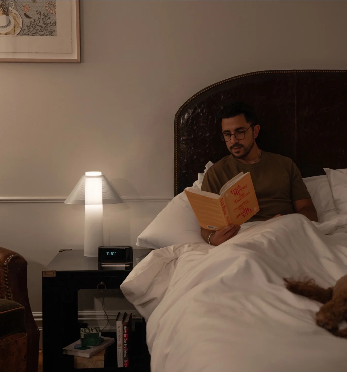 A person is sitting in bed reading a book titled "How to Disappear," with a Loftie alarm clock softly illuminating the room alongside the lamp on their nightstand.