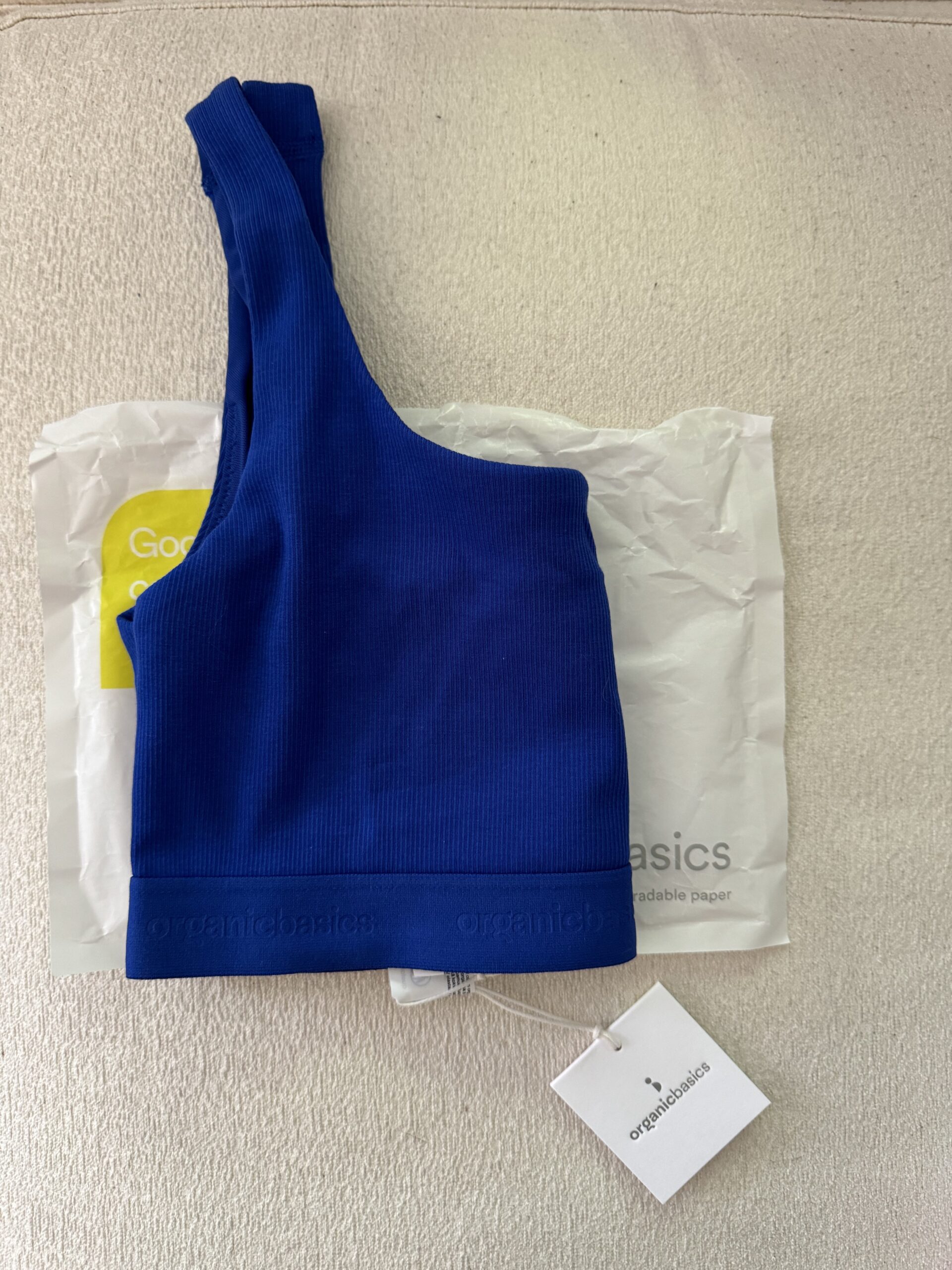 Blue one-shoulder top laid on packaging with a tag, on a textured beige surface.