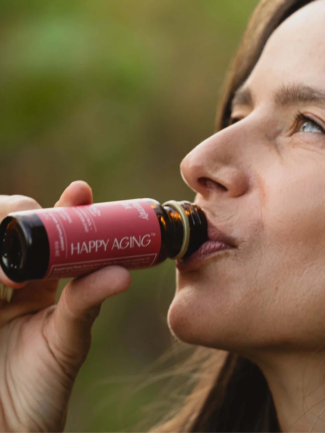 Person drinking from a small bottle labeled "Happy Aging" outdoors.