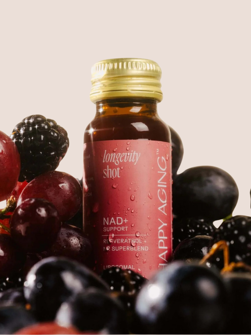 A bottle of "Longevity Shot" surrounded by assorted grapes and blackberries on a light background.