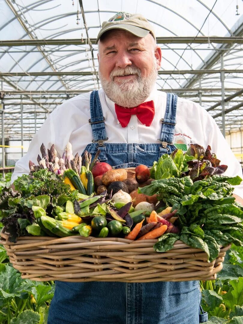 Person in overalls and a red bow tie holds a basket of assorted vegetables inside a greenhouse.