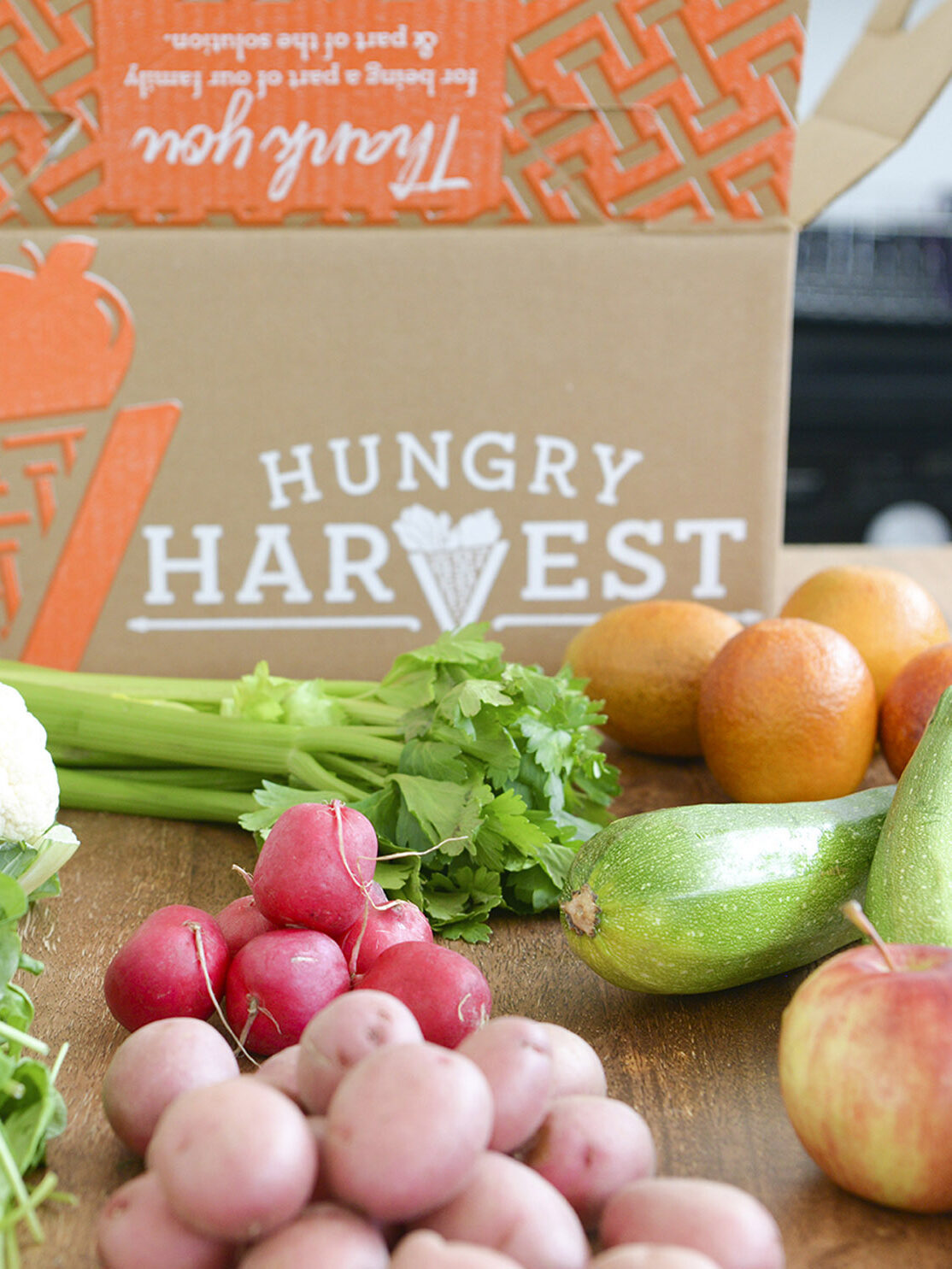 A box labeled "Hungry Harvest" surrounded by fresh produce including radishes, potatoes, cauliflower, celery, zucchini, oranges, and greens on a wooden surface.