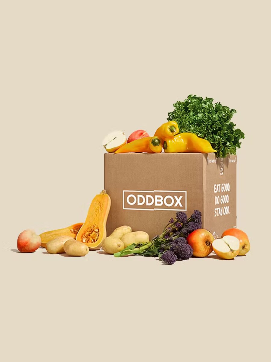 A cardboard box labeled "ODDBOX" is filled with various vegetables and fruits, including squash, apples, kale, peppers, potatoes, and plums, with some produce scattered around it.