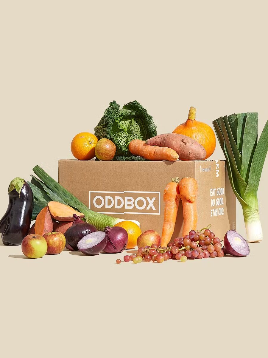 Box labeled "ODDBOX" surrounded by various fruits and vegetables, including eggplant, apples, onions, carrots, and leeks, on a neutral background.