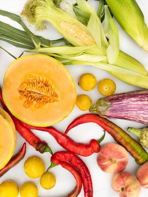 A variety of fruits and vegetables on a white background, including cantaloupe, corn, heirloom tomatoes, eggplants, peaches, hot peppers, and small yellow fruits.