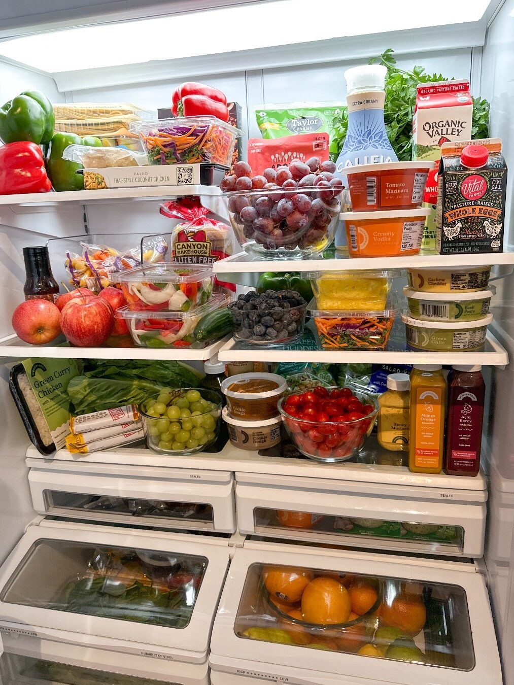 A well-organized refrigerator filled with various colorful fruits, vegetables, juice bottles, plant-based products, and packaged foods on neatly arranged shelves.