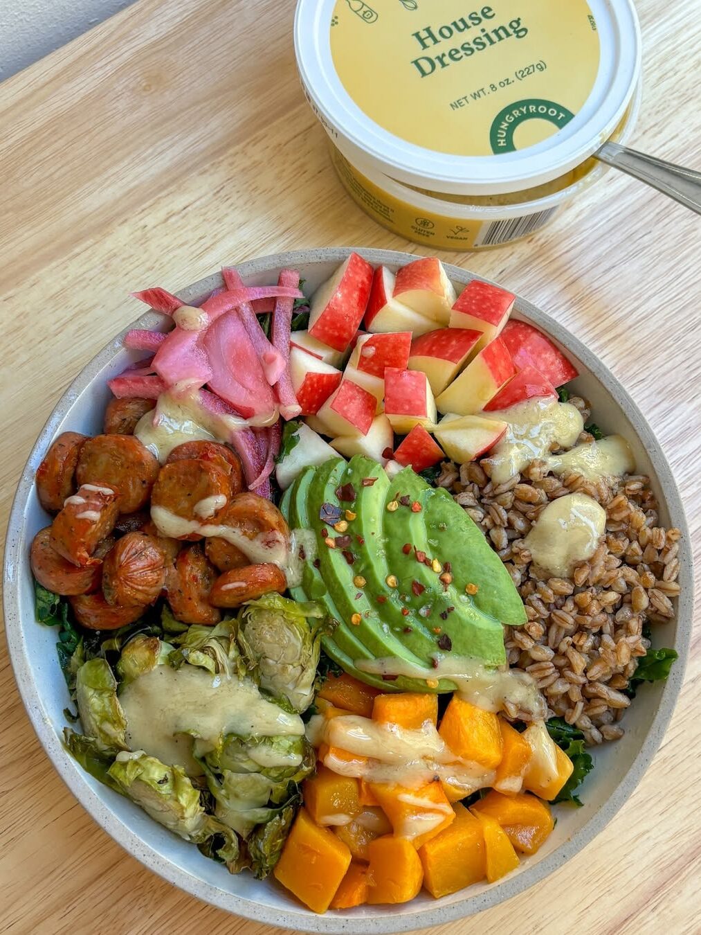 A bowl with avocado, grains, apples, Brussels sprouts, squash, pickled onions, and sausage, drizzled with dressing. Container of house dressing in the background.