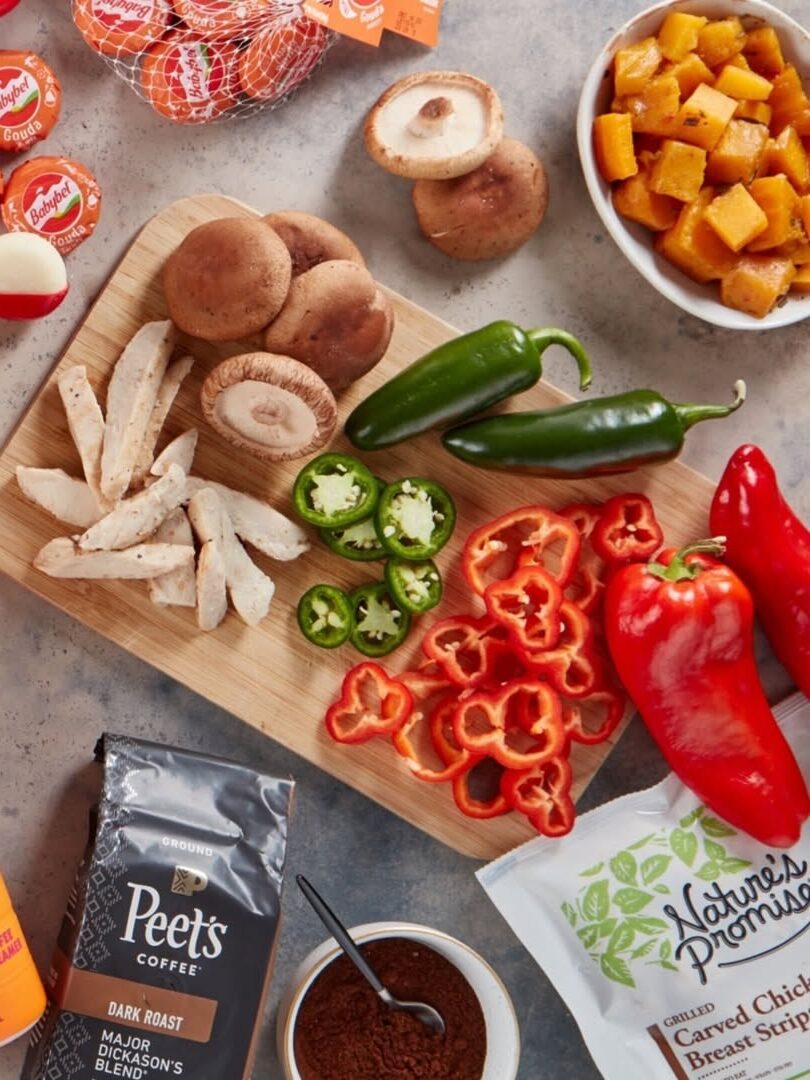 A variety of foods are arranged: cheese, iced coffee, mushrooms, peppers, pumpkin chunks, chicken strips, coffee beans, Dunkin' and Peet's coffee packs on a gray surface.