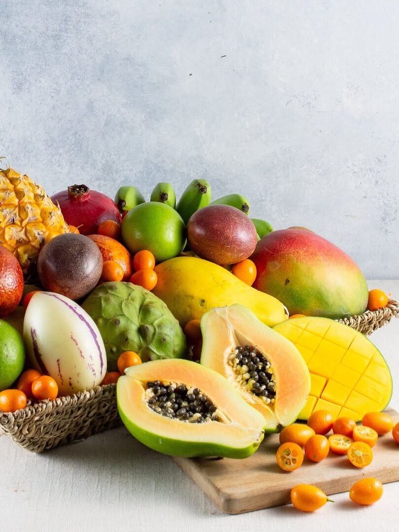 A variety of tropical fruits including pineapple, mango, papaya, bananas, and others are arranged in a basket. Sliced papaya and mango are on a wooden board in the foreground.