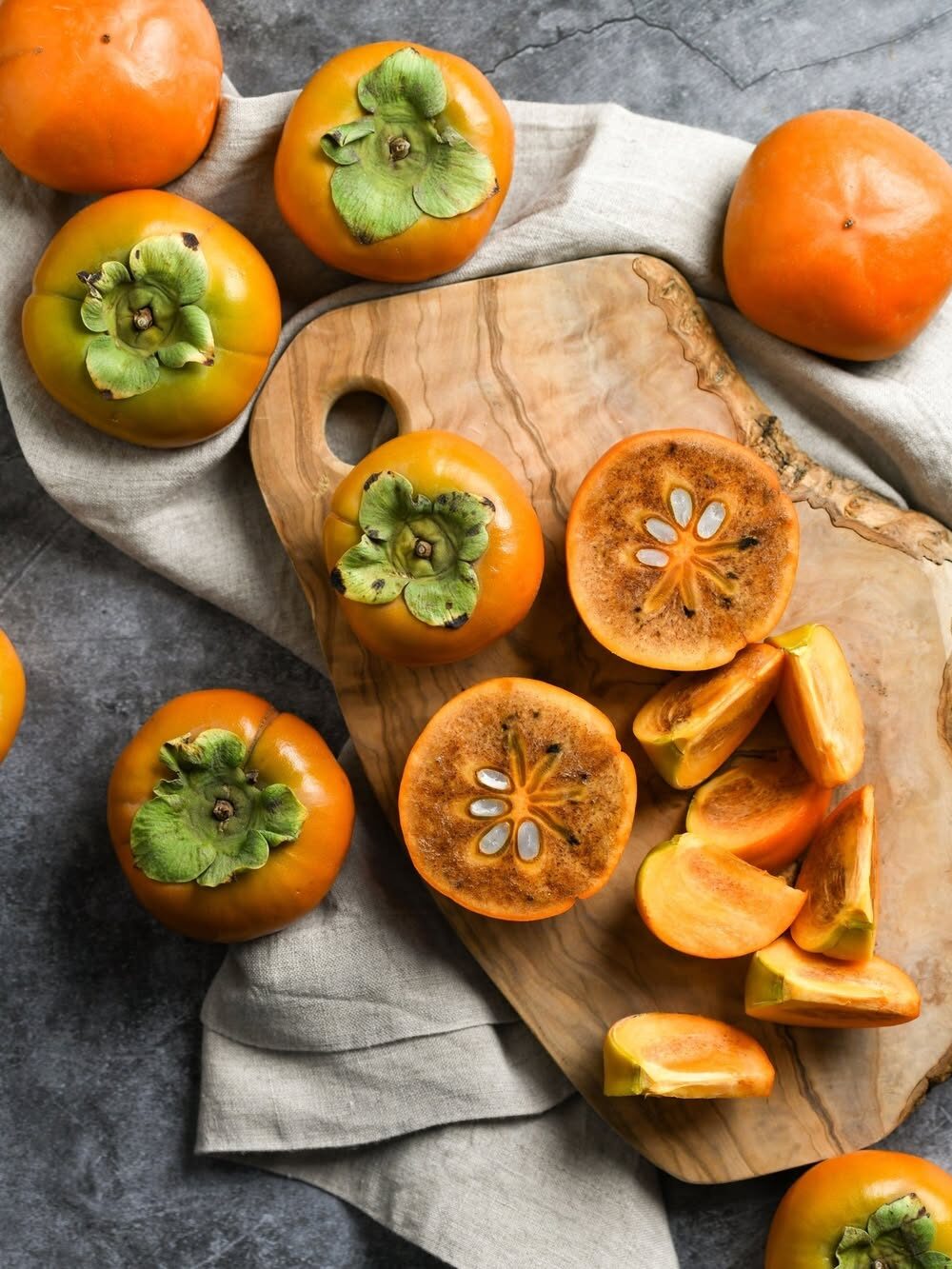 Persimmons, whole and sliced, are displayed on a wooden cutting board with a cloth underneath.