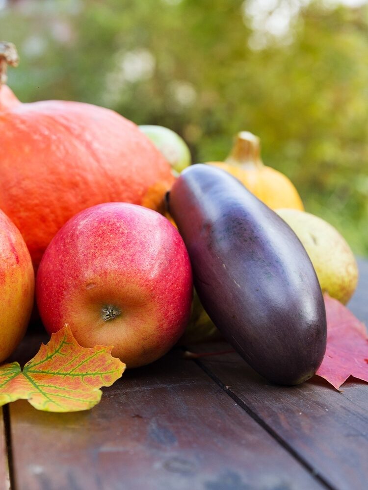 Colorful autumn harvest with a pumpkin, apples, eggplant, and leaves on a wooden table outdoors.