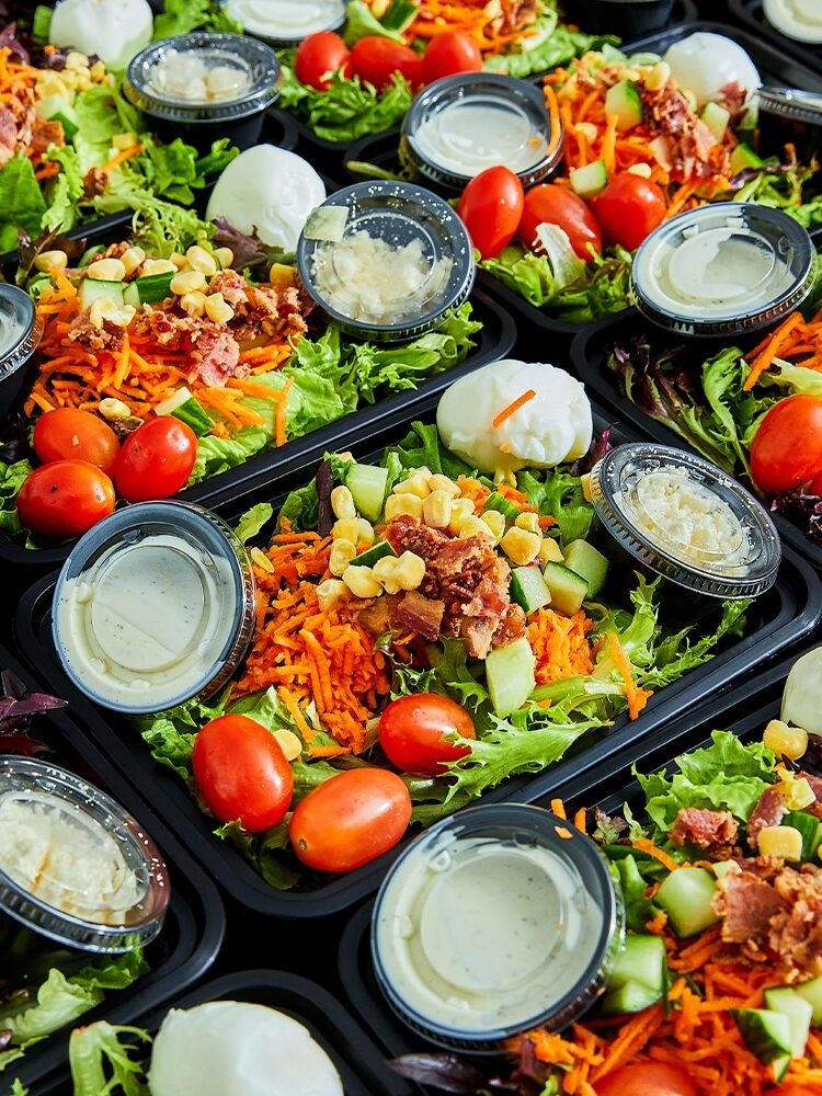 Rows of meal trays filled with salad, including lettuce, cherry tomatoes, shredded carrots, cucumbers, corn, bacon bits, and hard-boiled eggs. Small containers of dressing are included.