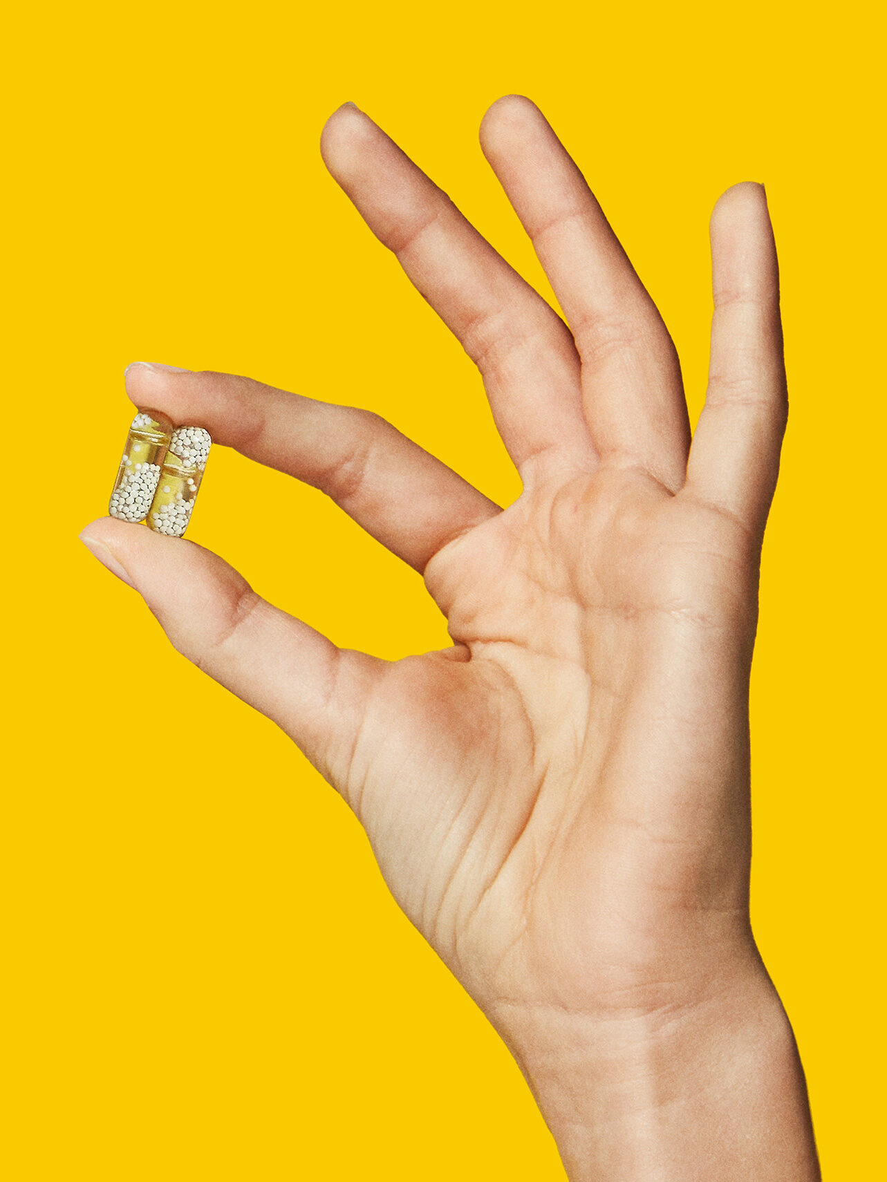 Hand holding two capsules filled with golden glitter against a solid yellow background.